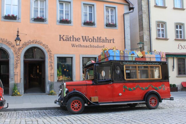 Lo storico autobus rosso e nero di Käthe Wohlfahrt, il Geschenkeexpress, parcheggiato sulla strada in ciottolato con il portapacchi sul tetto carico di pacchi regalo colorati con fiocchi dorati. Sullo sfondo la facciata color pesca del villaggio di Natale con finestre decorate da fioriere.